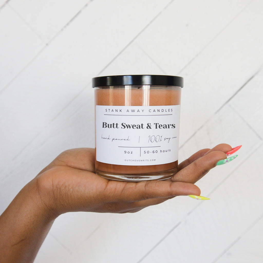Picture of a womans hand holding a Stank Away Butt Sweat and Tears brown sugar and fig scented candle on a wood background.