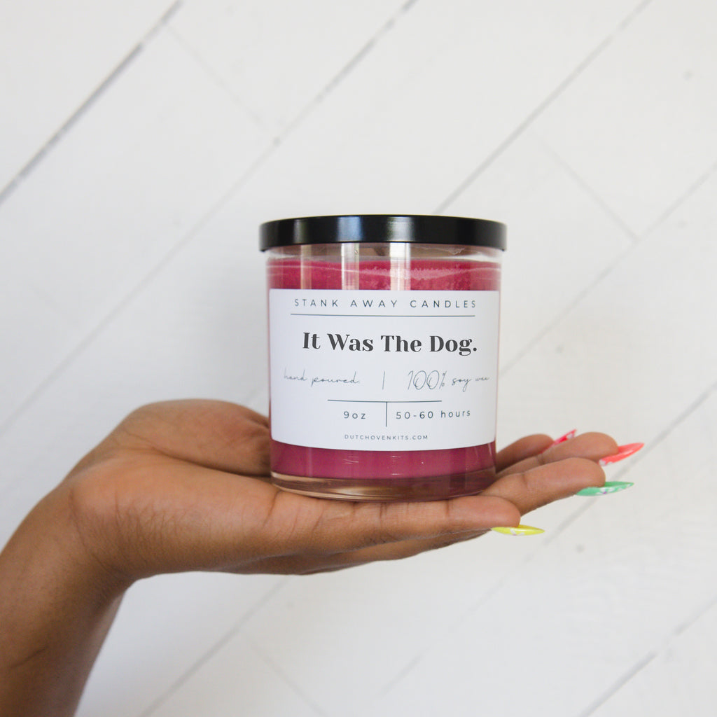 Picture of a womans hand holding a Stank Away It Was the Dog Wine Cellar scented candle on a wood background.
