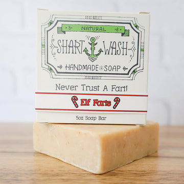 A box of shart wash elf farts scented soap on top of a tan soap bar on a wood table. Oatmeal spice scented holiday soap