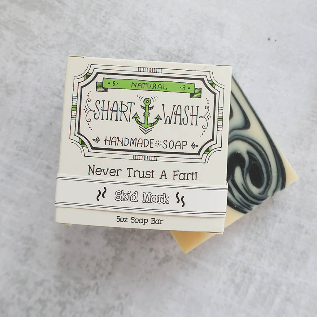Picture of a box of Shart Wash Natural Handmade Bar Soap, Skid Mark (Charcoal and Aloe) scent sitting on a black and white bar of soap with a wood background