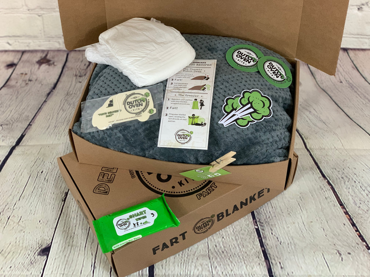 picture of a gray blanket in a box with an adult diaper, air freshener, pack of shart wipes, fart recipe card and stickers.