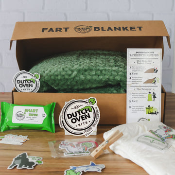 A green deluxe dutch oven kits gift box on a wood table with an adult diaper, pack of shart wipes, car air freshener, stickers, clothes pin and fart recipe card. Fart Blanket