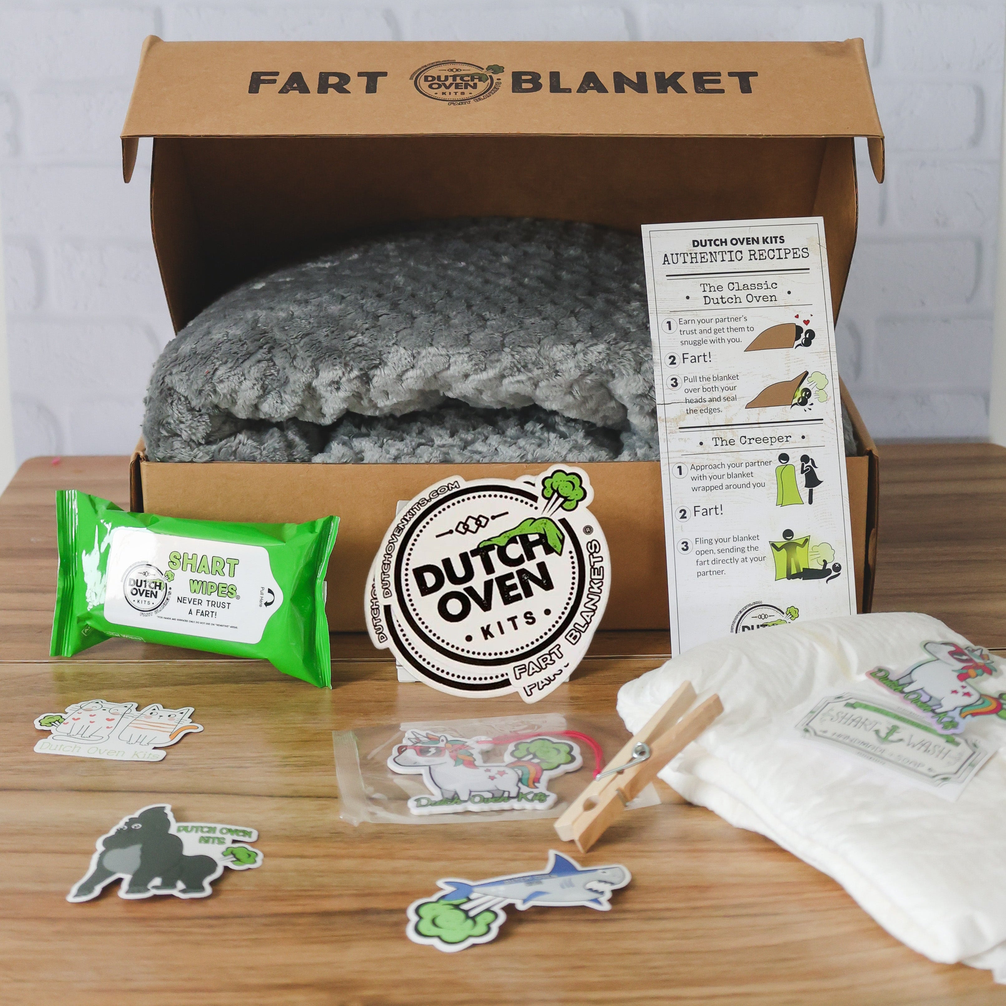 A gray deluxe dutch oven kits gift box on a wood table with an adult diaper, pack of shart wipes, car air freshener, stickers, clothes pin and fart recipe card. Fart Blanket