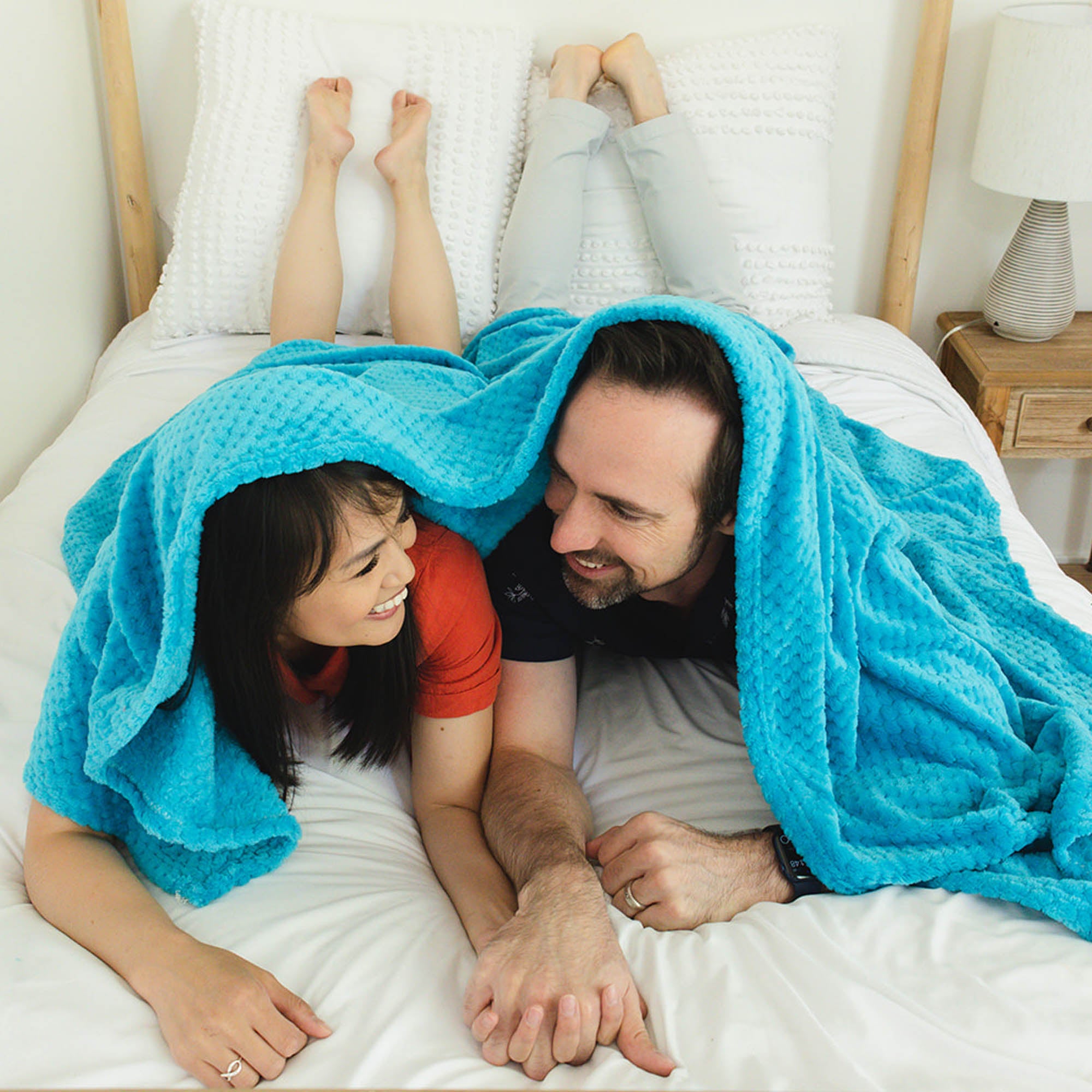 a smiling couple holds hands on a bed covered by  a blue blanket.
