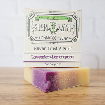 Picture of a box of Shart Wash Natural Handmade Bar Soap Lavender Lemongrass scent sitting on a purple and white bar of soap with a wood background