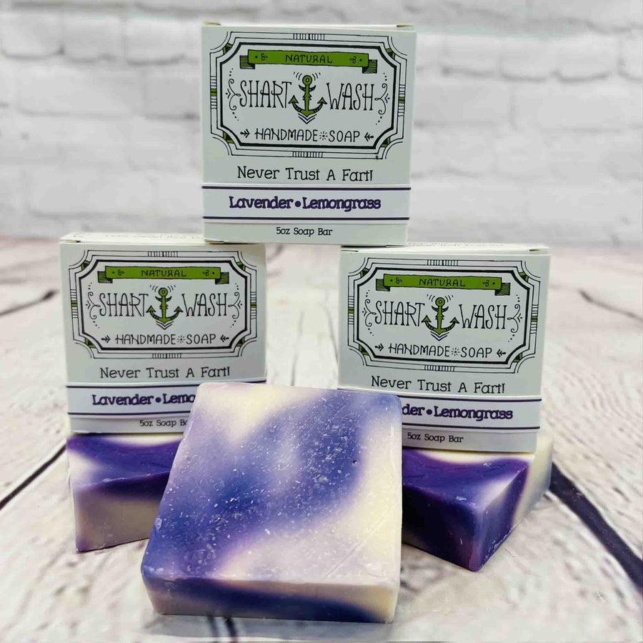 Picture of 3 boxes of Shart Wash Natural Handmade Bar Soap Lavender Lemongrass scent sitting on two purple and white bars of soap with a wood background